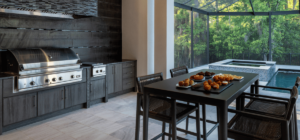 An elegant outdoor kitchen featuring sleek stainless steel appliances, dark cabinetry, and a modern dining table set. The space overlooks a luxurious pool and spa surrounded by lush greenery, creating a serene and inviting environment.