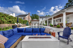 Luxury backyard outdoor living space with a modern pool, fire pit, blue sectional seating, and a covered patio featuring holiday decorations, lounge chairs, and a flat-screen TV.