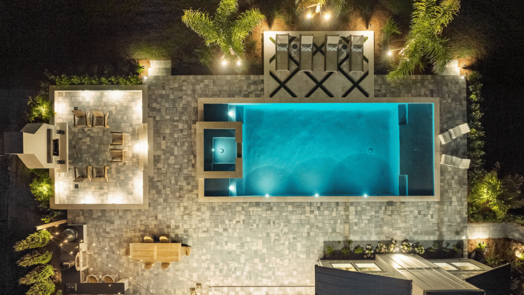 Aerial view of a luxurious backyard swimming pool with integrated spa and surrounding patio areas illuminated at night, showcasing elegant landscaping and outdoor furniture.